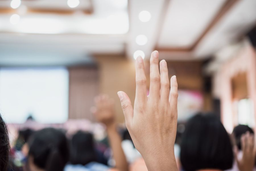 Eine Hand wird in einem Raum voller Menschen hochgehoben, als ob sie eine Frage stellen oder sich melden möchte.