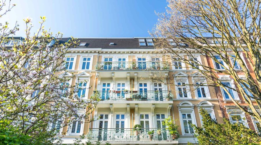 Ein historisches Mehrfamilienhaus mit Balkonen und blühenden Bäumen davor unter einem klaren blauen Himmel.