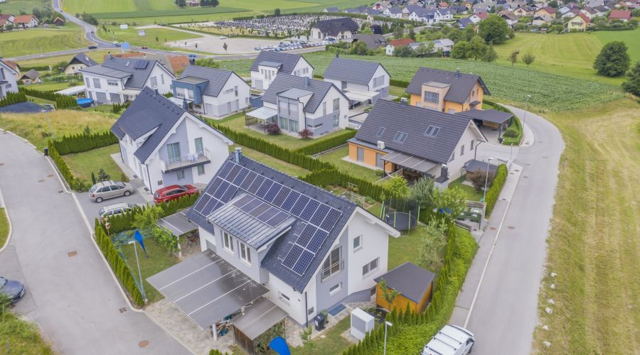 Eine moderne Wohnsiedlung mit Einfamilienhäusern, teilweise mit Solarpanels auf den Dächern. Die Umgebung ist ländlich geprägt mit grünen Feldern und Hügeln. Die Straßenführung ist kurvig und sauber, eingebettet in gepflegte Gärten.