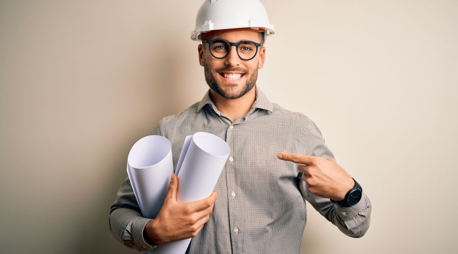 Ein lächelnder Architekt mit einem Bauhelm hält gerollte Baupläne und zeigt mit dem Finger darauf.