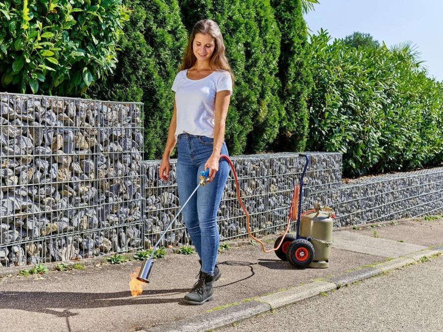 Eine Frau benutzt ein Abflammgerät, um Unkraut auf einem Gehweg neben einer Gabionenwand zu entfernen.