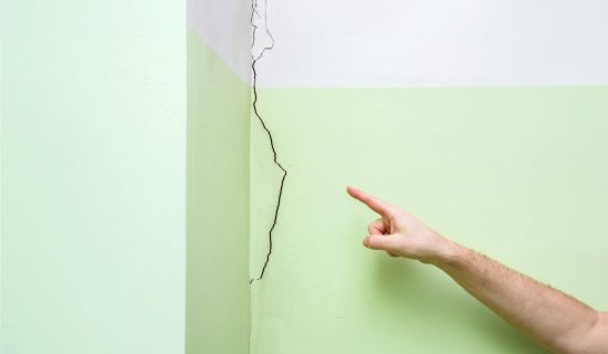 Das Bild zeigt eine grüne Wand in der Ecke eines Raumes mit einem Riss, der von oben bis unten verläuft. Ein Finger zeigt auf den Riss, der gut sichtbar ist.