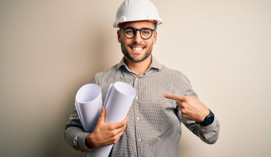 Ein lächelnder Architekt mit einem Bauhelm hält gerollte Baupläne und zeigt mit dem Finger darauf.