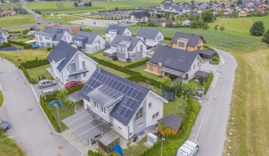 Eine moderne Wohnsiedlung mit Einfamilienhäusern, teilweise mit Solarpanels auf den Dächern. Die Umgebung ist ländlich geprägt mit grünen Feldern und Hügeln. Die Straßenführung ist kurvig und sauber, eingebettet in gepflegte Gärten.