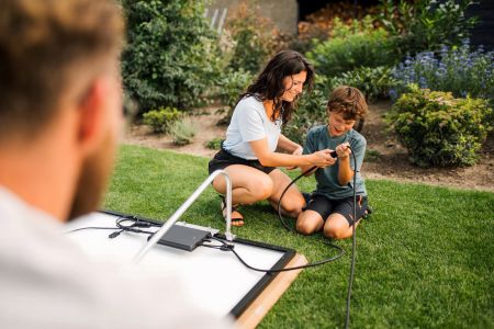 Frau und Kind installieren ein Solarpanel im Garten, während ein Mann im Vordergrund zuschaut.