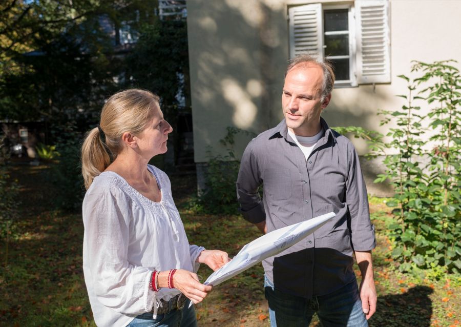 Zwei Personen stehen in einem Garten vor einem Haus und unterhalten sich, während eine Person Baupläne in den Händen hält.