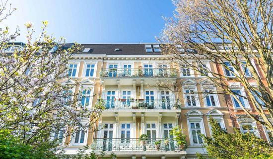 Ein historisches Mehrfamilienhaus mit Balkonen und blühenden Bäumen davor unter einem klaren blauen Himmel.