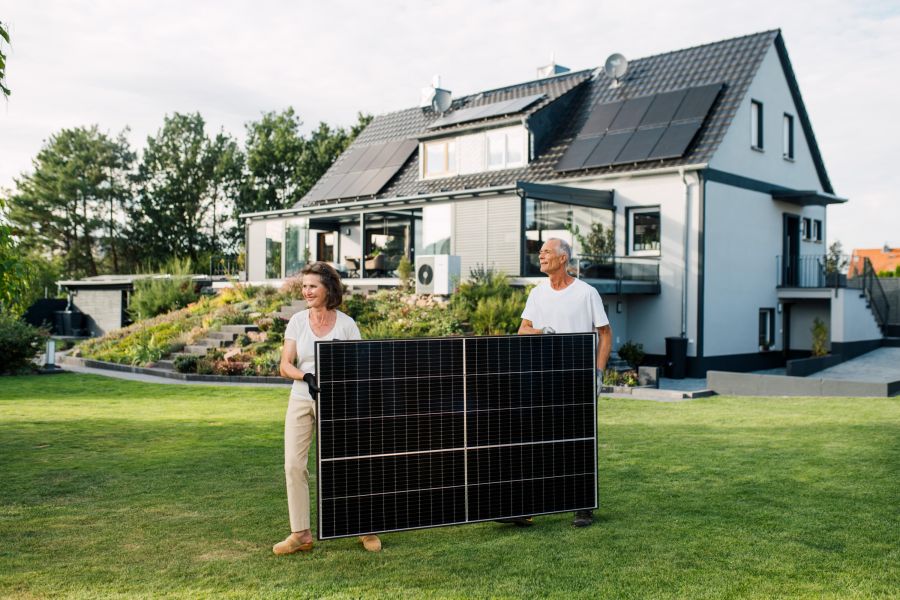 Ein älteres Paar hält ein Solarmodul auf dem Rasen vor einem modernen Haus mit bereits installierten Solarpanels auf dem Dach. Der gepflegte Garten und die Terrassenanlage im Hintergrund vermitteln eine nachhaltige und umweltbewusste Lebensweise. Das son