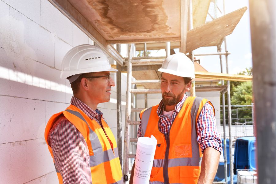 Zwei Männer in Bauhelmen und orangenen Warnwesten stehen auf einer Baustelle und besprechen den Bauplan.