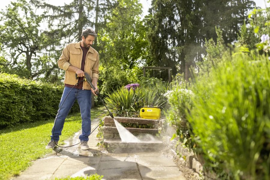 Ein Mann reinigt eine Terrasse im Garten mit einem Hochdruckreiniger, umgeben von Pflanzen und Rasen.