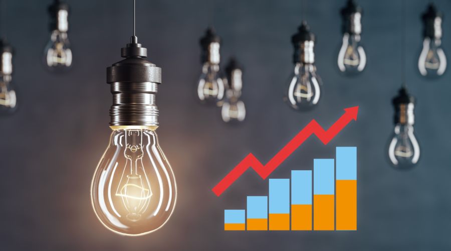 Das Bild zeigt eine leuchtende Glühbirne im Vordergrund, umgeben von weiteren Glühbirnen im Hintergrund, sowie einem Diagramm mit aufsteigenden Balken in Blau und Orange und einem roten Pfeil, der den Wachstumstrend symbolisiert.