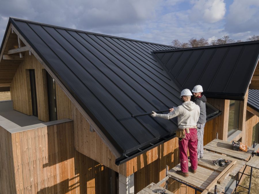 Zwei Arbeiter mit Schutzhelmen installieren eine schwarze Metall-Dacheindeckung auf einem modernen Holzhaus. Das Dach hat eine klare, minimalistische Architektur, während der Himmel im Hintergrund von Wolken bedeckt ist.