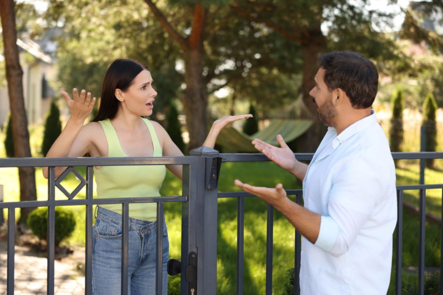 Ein Mann und eine Frau diskutieren lebhaft über einem Metallzaun in einem grünen Garten.