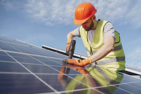 Ein Techniker in reflektierender Sicherheitsweste und orangefarbenem Schutzhelm arbeitet mit einem Elektrowerkzeug an einer Solarpanel-Installation. Der sonnige Himmel im Hintergrund unterstreicht die nachhaltige Energieerzeugung. Die Solarpanels reflekti