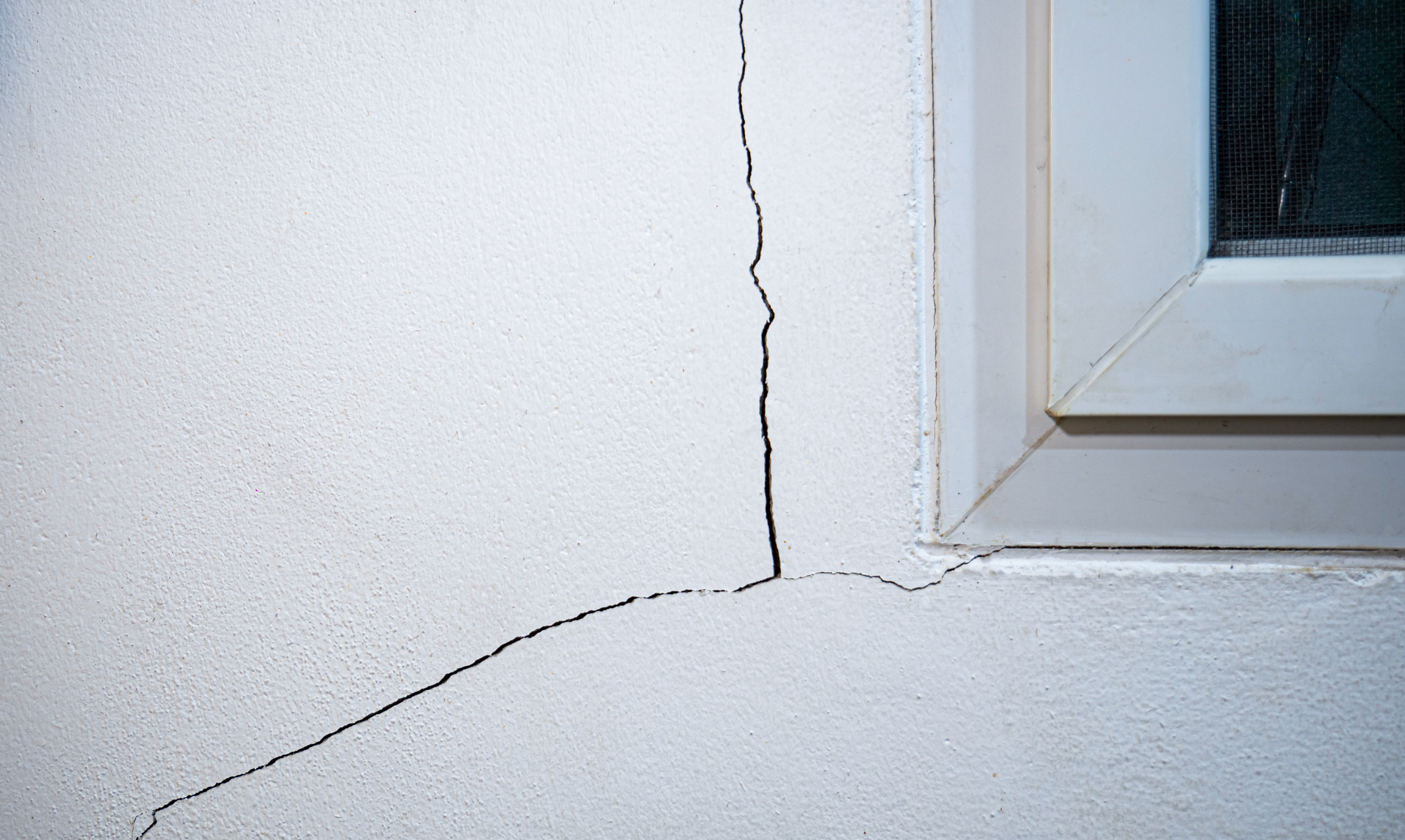 Das Bild zeigt eine weiße Wand mit einem schmalen, diagonal verlaufenden Riss. Oben rechts ist ein weißer Fensterrahmen sichtbar, der von der Wand abgesetzt ist.